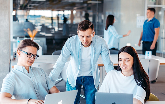 Two people sitting down on laptops and man standing behind them leaning down