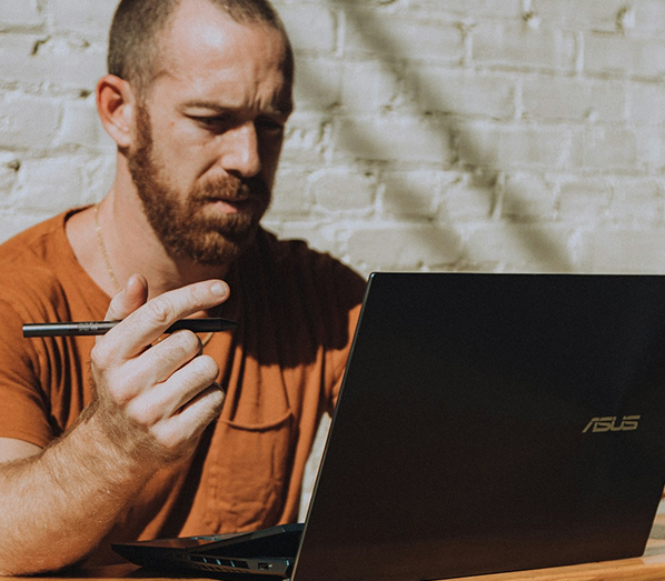 Man with beard holding pen looking at laptop
